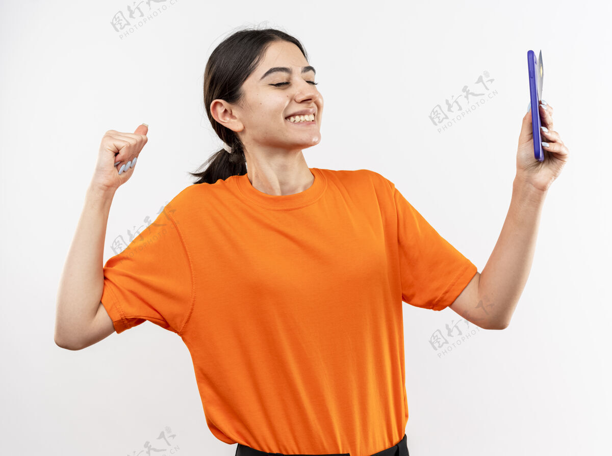手机身穿橙色t恤的年轻女孩手持智能手机握紧拳头 在白色背景上快乐而兴奋地站着T恤握着拳头