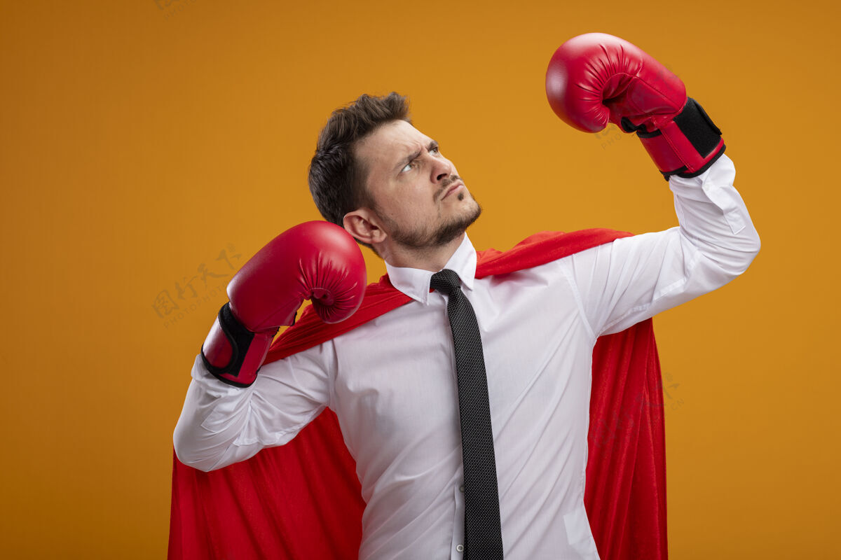 严肃严肃自信的超级英雄商人穿着红色斗篷 戴着拳击手套 站在橙色背景下举手展示力量和勇气力量提高显示