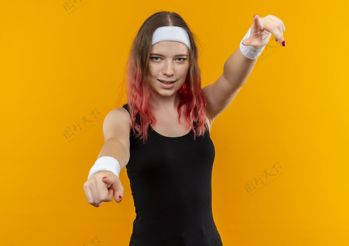 运动员身着运动服的年轻健身女士 带着自信的微笑 手指着站在橙色墙上的摄像机手指女性人