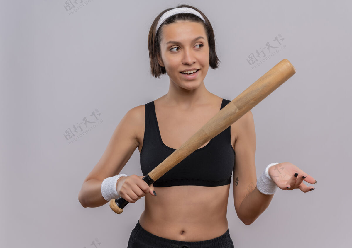 运动装身穿运动服 手持棒球棒 脸上带着自信微笑的年轻健身女士站在白色的墙上信心年轻棒球