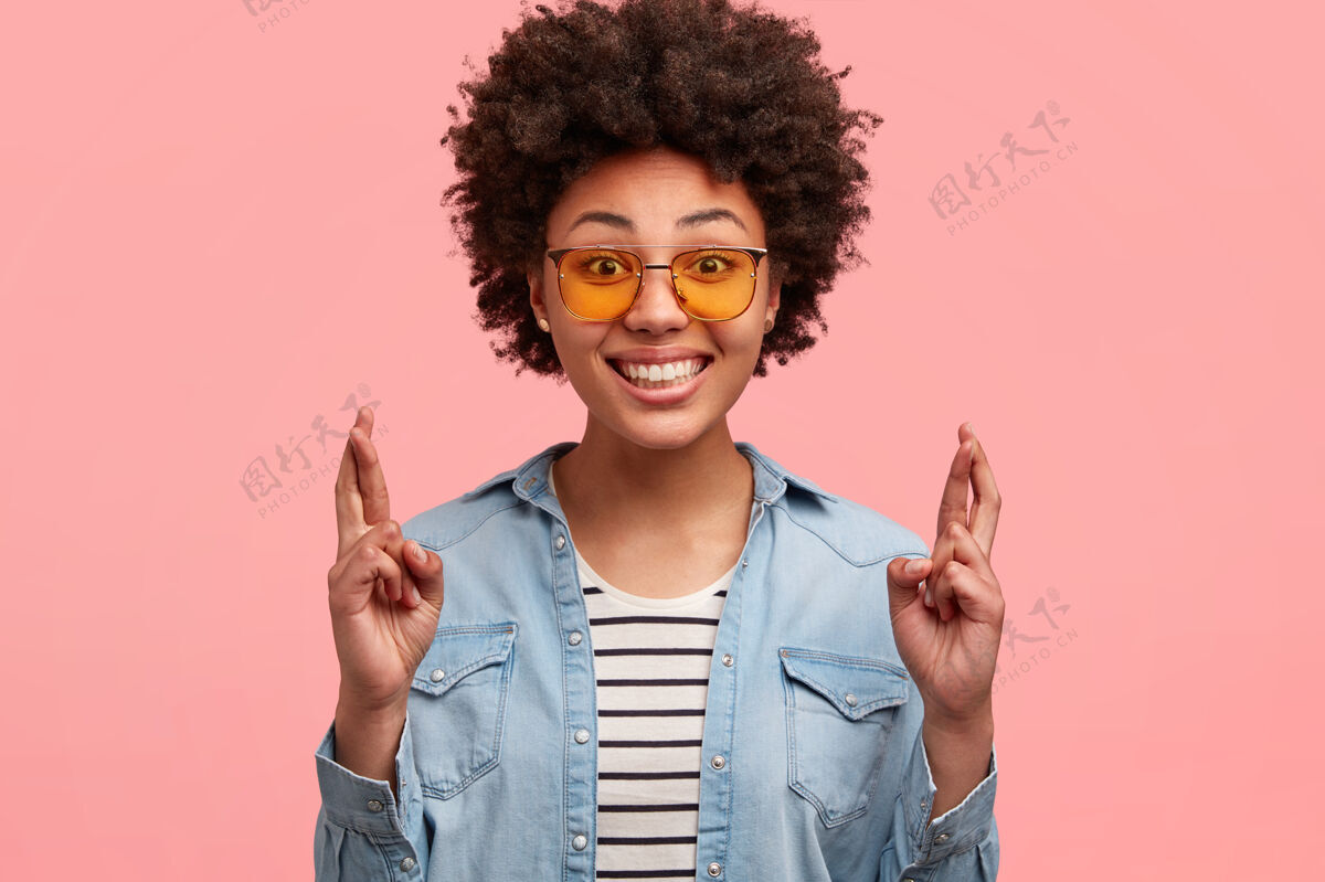 成人快乐的年轻美国黑人妇女渴望梦想成真 双手交叉许愿 戴着时髦的墨镜 笑容灿烂 露出完美的牙齿 在粉红色的墙上做模特手指非洲女人