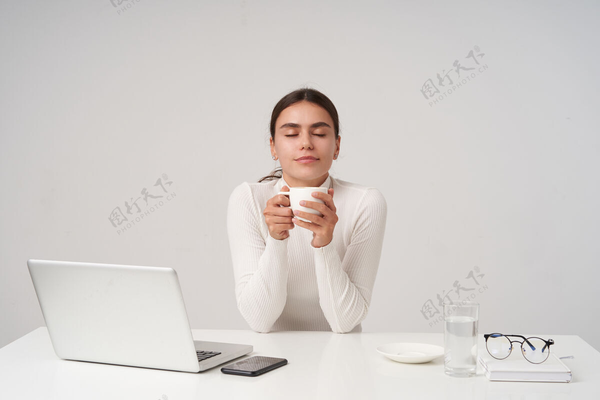 发型喜欢年轻漂亮的黑发女人 扎着马尾辫 喝着咖啡休息 拿着白色陶瓷杯 闭着眼睛愉快地微笑 坐在白色的墙上女士眼镜笔记本电脑