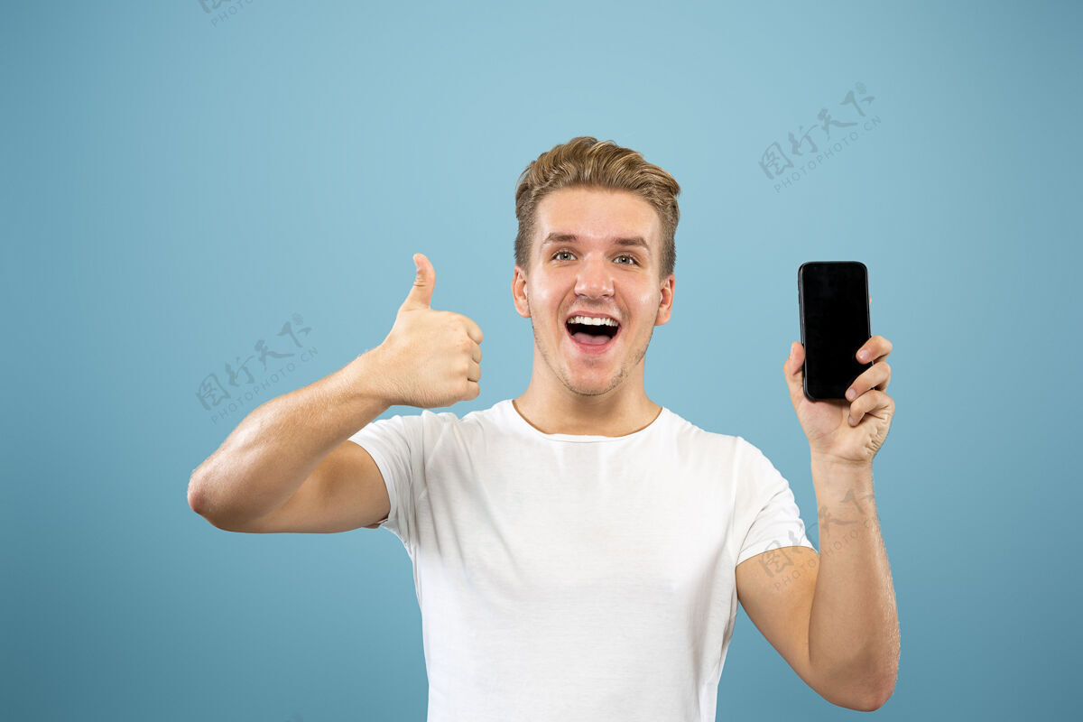 衬衫在蓝色工作室背景上的白人年轻人半身肖像穿着衬衫的漂亮男模人类情感的概念 面部表情 销售 广告显示电话屏幕 付款 投注面部商人表情