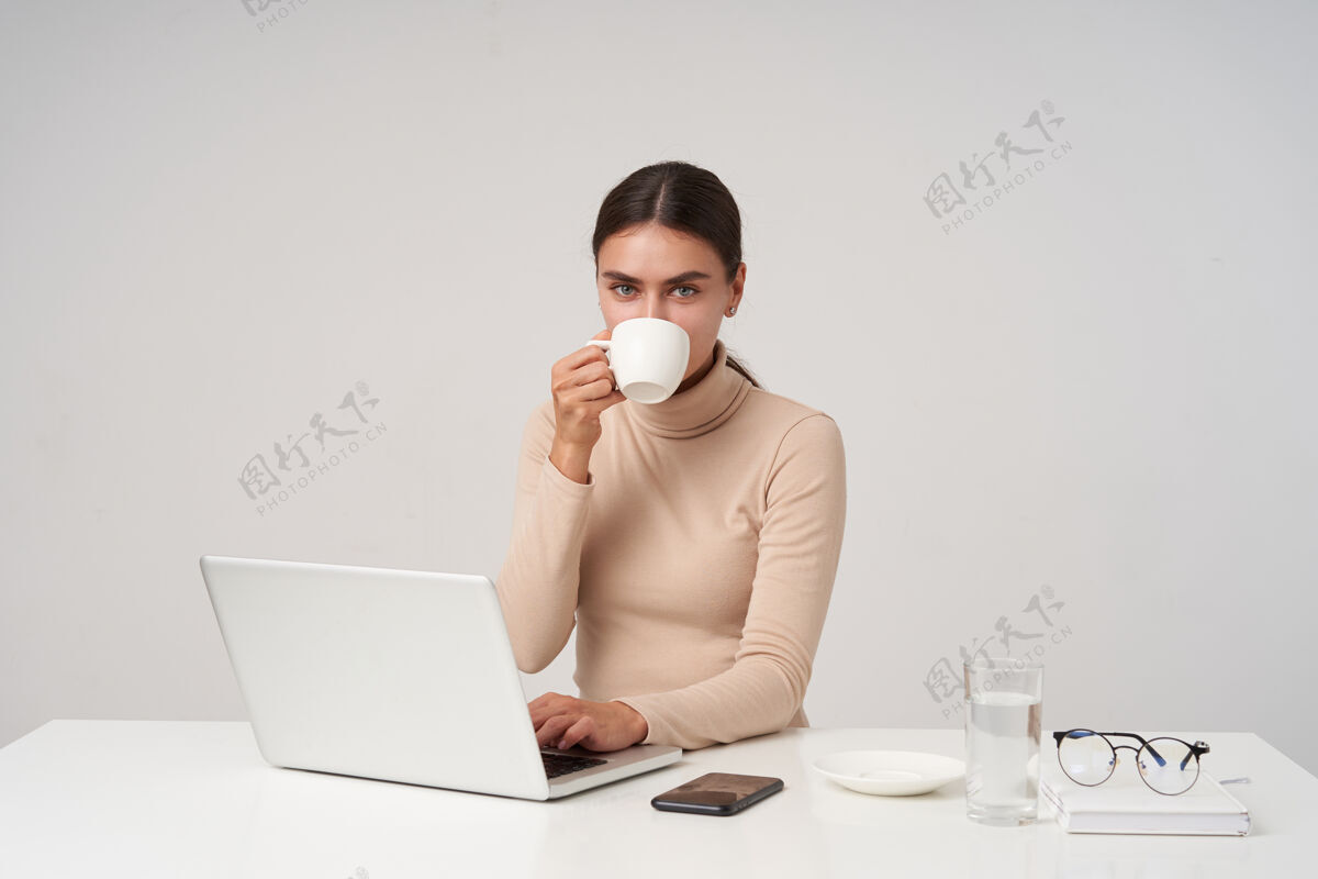 发型迷人的年轻蓝眼睛黑发女子 穿着米色马球衫 一边喝咖啡一边在键盘上打字 坐在白色的墙上积极地看着头发穿着工作