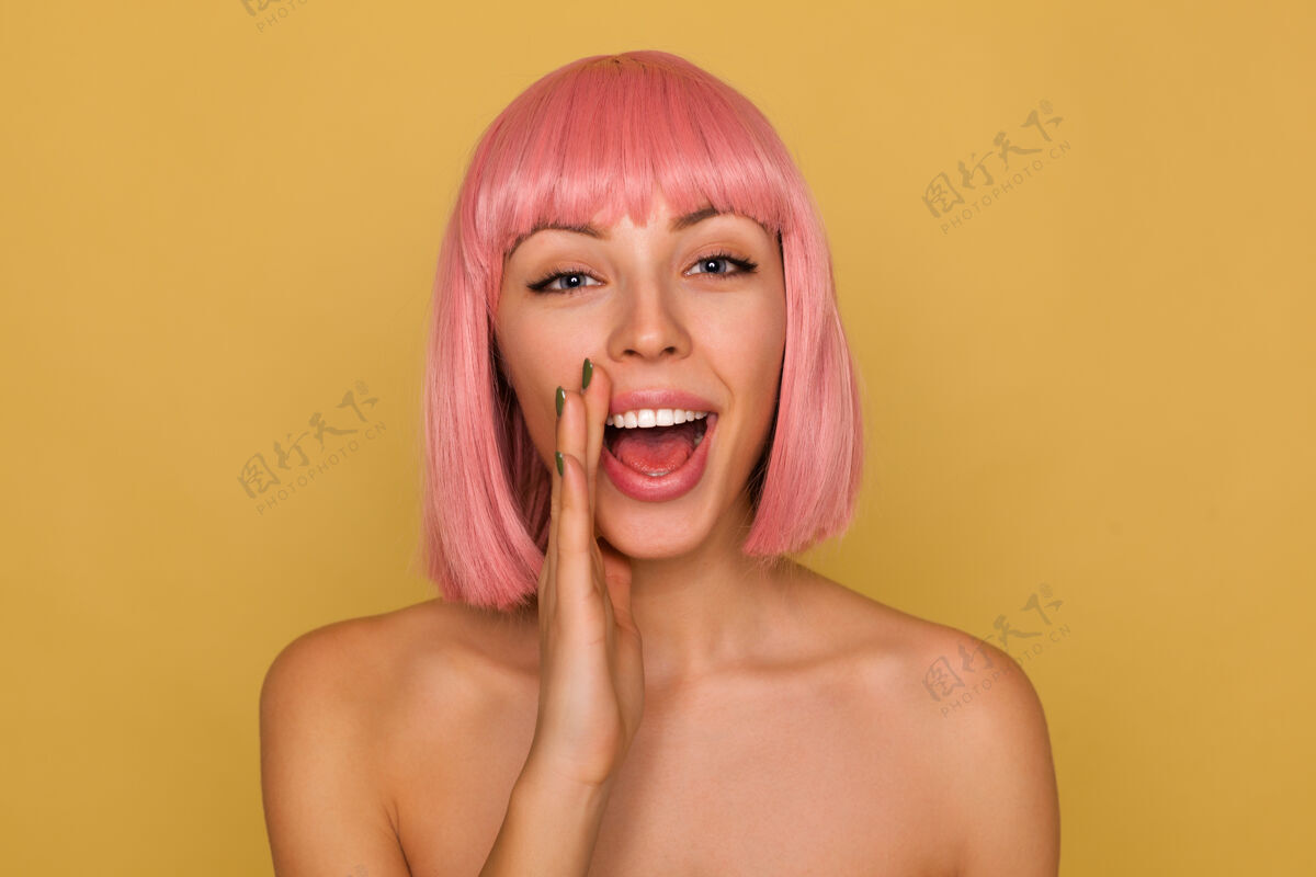模特特写镜头：兴奋的年轻可爱的蓝眼睛女士 粉色的鲍勃发型 举起手捂着脸喊着什么 赤裸着肩膀站在芥末墙上动画自然表情