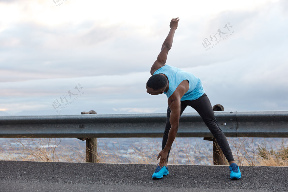 乡村水平拍摄的深色皮肤的男人靠在脚上 做伸展运动 有肌肉的体形 站在浅蓝天空的沥青路面上 为你的广告内容 文字复制空间马拉松民族运动员
