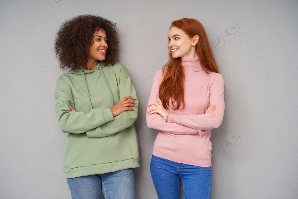 年轻人室内拍摄的可爱开朗的年轻女性 随意的发型交叉双手 站在灰色的墙上 积极地看着对方 愉快地微笑Foxy模特女人