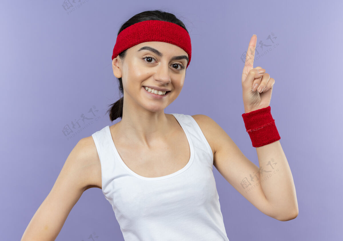 运动身穿运动服的年轻健身女士 头箍朝上 食指微笑 自信地站在紫色的墙上头带运动员健身