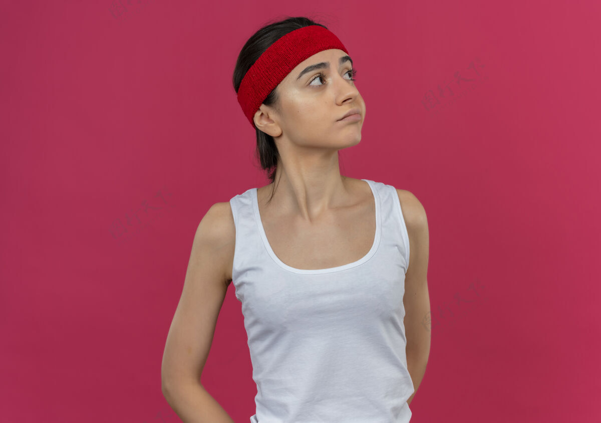 运动戴着头巾 穿着运动服的年轻健身女士 站在粉红色的墙上 带着怀疑的表情向一旁望去姿势怀疑运动员