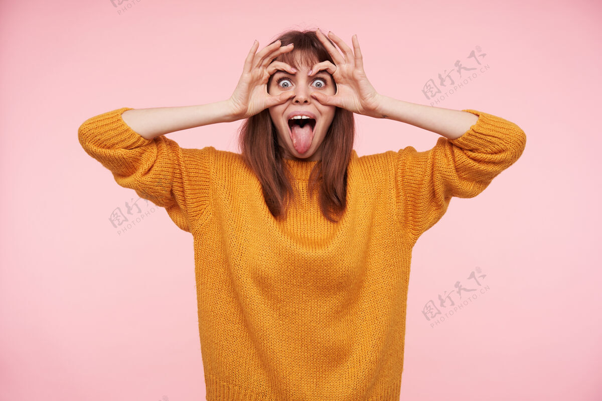 穆阿身着芥末色羊毛衫站在粉色墙壁上 年轻快乐的黑发女士伸出舌头 双手放在眼睛上的画像欢呼表情女性