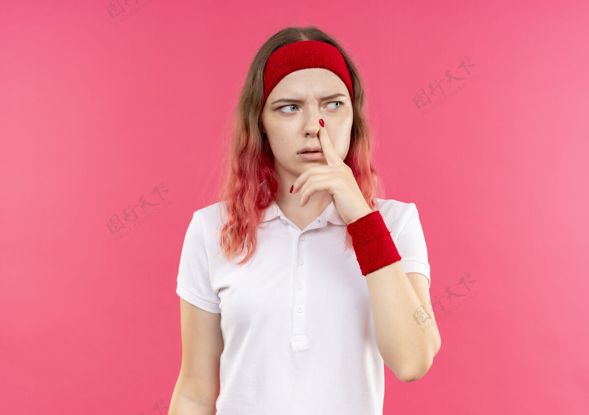 运动戴着头巾的年轻运动女郎站在粉红色的墙上 一边看一边抚摸着鼻子 脸上带着沉思的表情女人市民运动员
