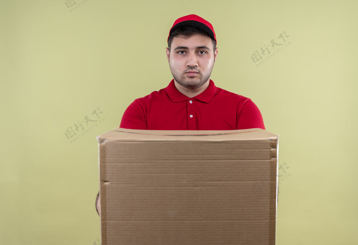 人年轻的送货员穿着红色制服 戴着帽子 一副严肃的面孔站在绿色的墙上摆姿势拿着送货