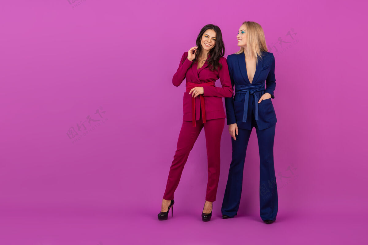 华丽时尚风格两位微笑迷人的女士在紫罗兰色的墙壁上穿着紫色和蓝色的时尚多彩的晚礼服 春天的时尚潮流穿着时尚衣服