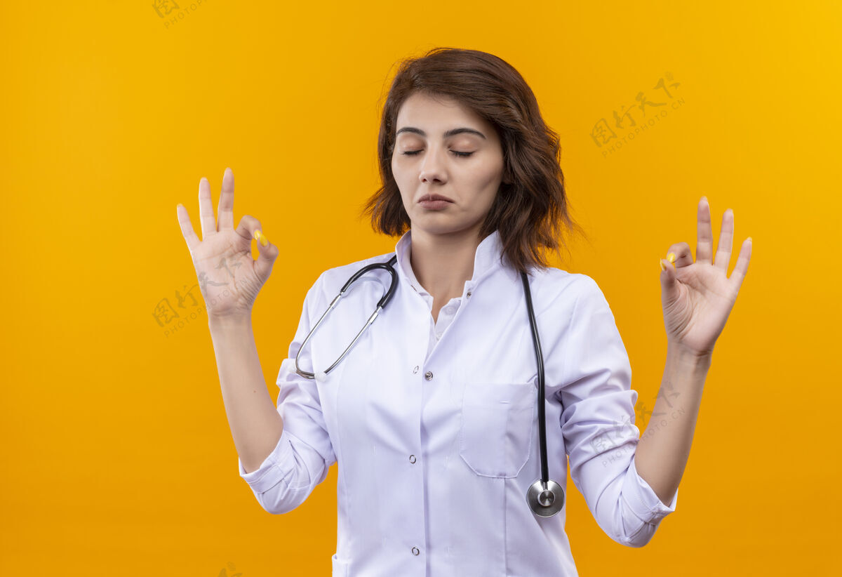 听诊器年轻的女医生 穿着白大褂 戴着听诊器 闭着眼睛放松 做着冥想的手势 手指站在橙色的墙上手势医生手指