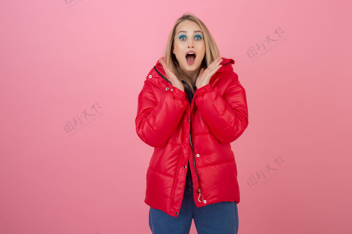 时尚金发碧眼的活跃女性穿着色彩鲜艳的红色冬季羽绒服在粉色墙面上摆造型 尽情玩乐 温暖的外套时尚潮流 惊喜震惊的表情颜色服装夹克