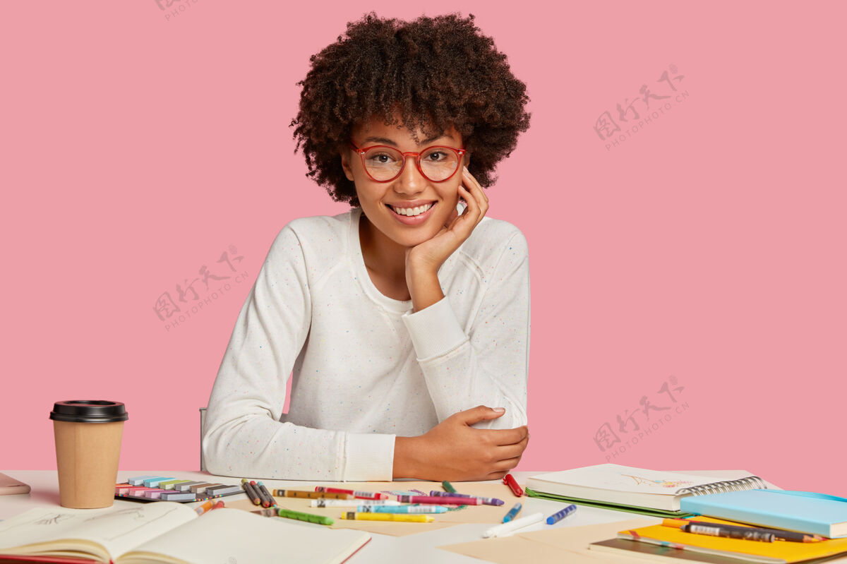 工作区令人愉快的黑人发型 手贴在脸颊上 戴着眼镜 穿白色衣服 在笔记本上画草图 模特在粉色墙上的桌面上工作场所办公桌饮用