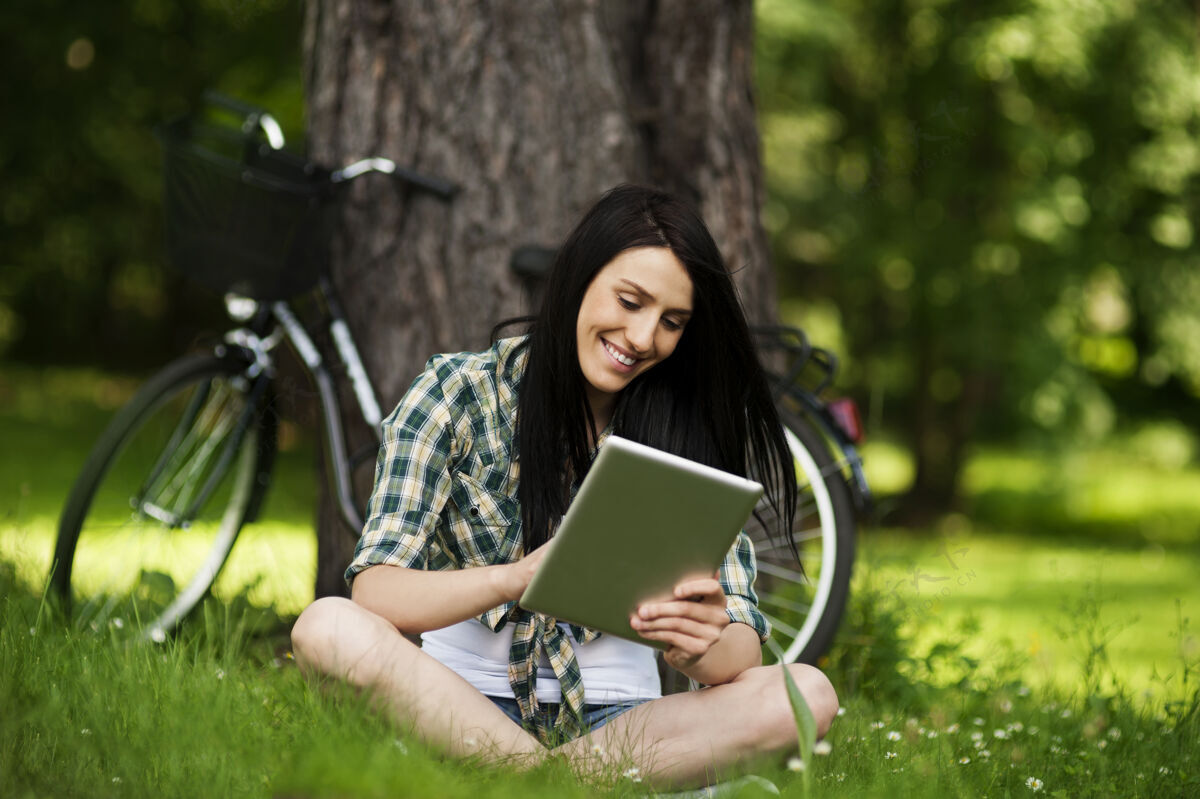 年轻人美丽的年轻女子在户外使用数码平板电脑自行车草地树