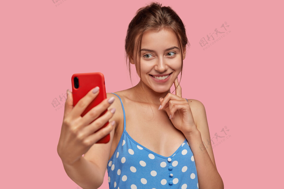 微笑积极的年轻白种女人用现代手机拍照 脸上带着温柔的微笑 穿着蓝色的波尔卡裙 在粉色的墙上做模特漂亮的女人摆出自拍的姿势青少年自拍视频通话