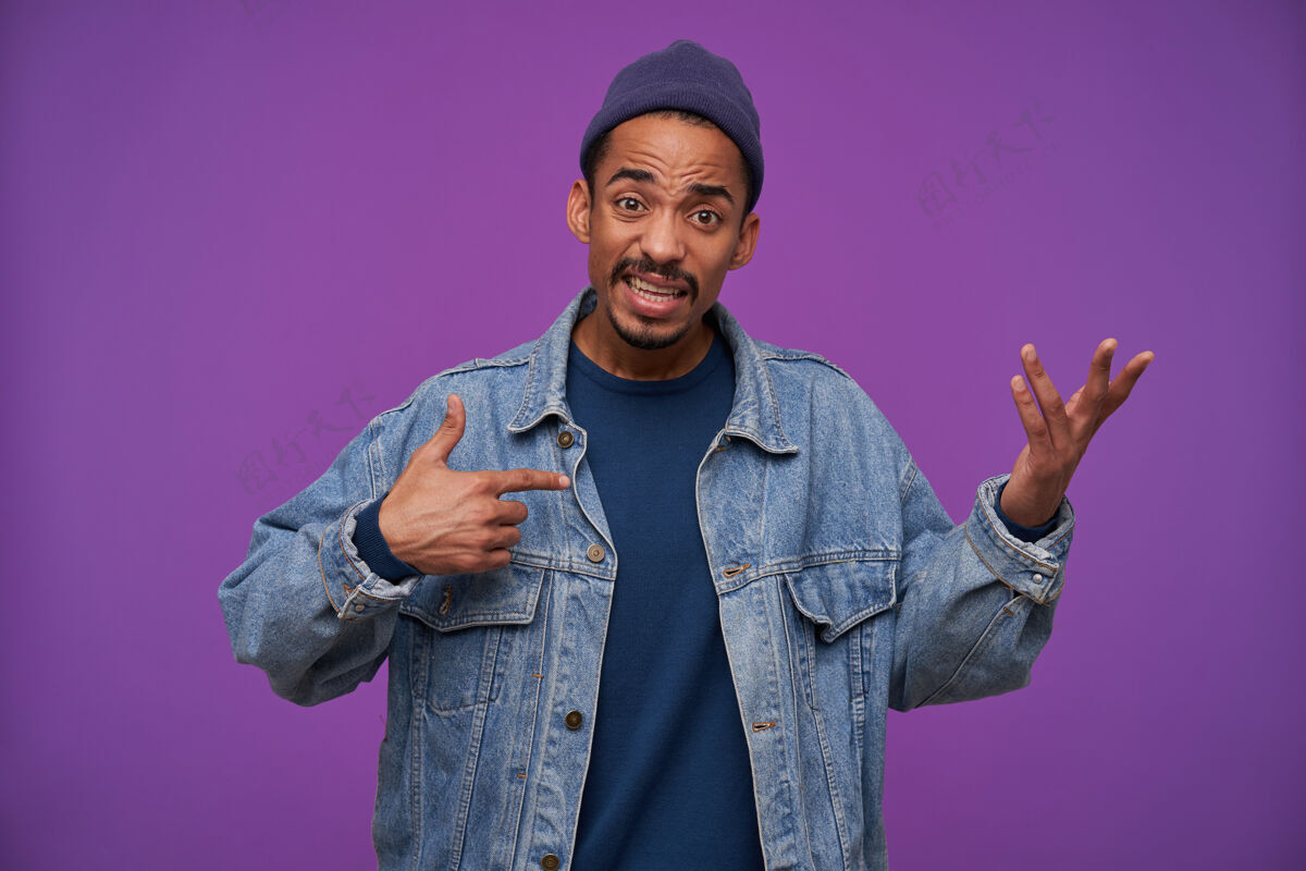 20岁困惑的年轻黑皮肤 胡子 深色头发的男性皱着额头 抬起迷茫的手一边看 穿着便服越过紫色的墙壁年轻肖像帽子