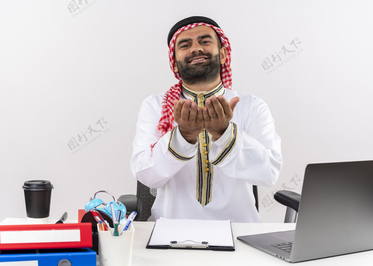 笔记本电脑身着传统服装的阿拉伯商人坐在桌旁 手拿笔记本电脑 在办公室里工作要钱拿问电脑