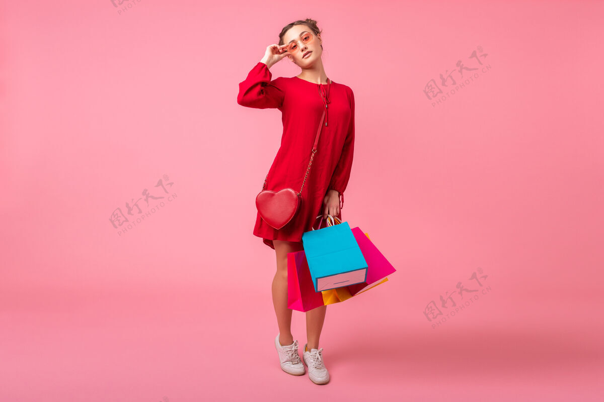 颜色迷人快乐微笑的时尚女性购物狂身着红色新潮连衣裙手持五颜六色的购物袋贴在粉色的墙上 销售兴奋 春夏时尚潮流休闲衣服购买
