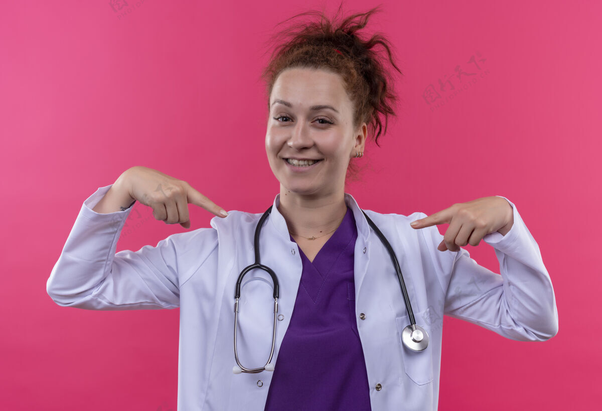 听诊器年轻的女医生穿着白大褂 用听诊器指着自己满意地站在粉红色的墙上医生她自己自我满足
