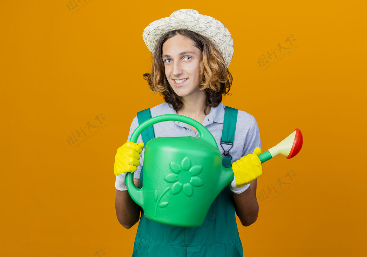 男人年轻的园丁戴着橡胶手套 穿着连体衣 戴着帽子 手里拿着浇水罐花园手套连身衣