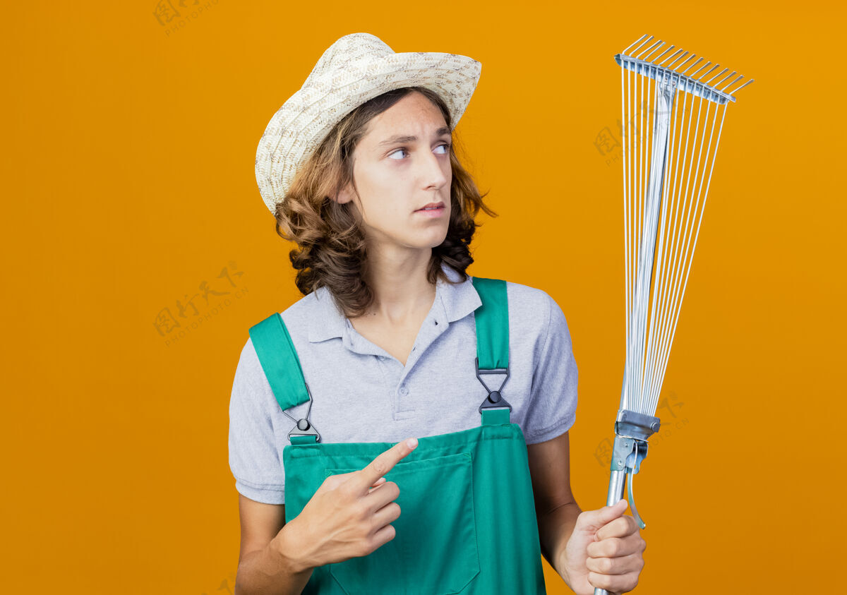 年轻年轻的园丁穿着连体衣戴着帽子拿着耙子用食指指着脸男人抱着