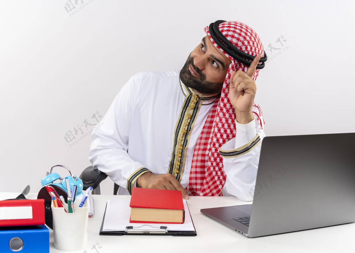 笔记本电脑身着传统服装的阿拉伯商人坐在桌子旁 手持笔记本电脑 一边看着一边 手指朝上 在办公室工作穿坐商务人士