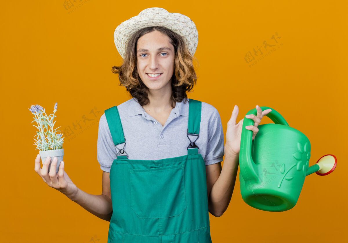 举行年轻的园丁 穿着连体衣 戴着帽子 手里拿着水罐和盆栽植物微笑连身衣年轻人