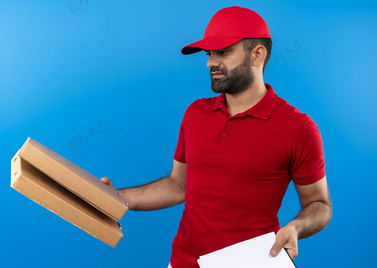 帽子一个留着胡须 身穿红色制服 头戴鸭舌帽的送货员站在蓝色的墙上 手里拿着披萨盒和剪贴板 空白的页面看上去很困惑 有疑问制服胡子有