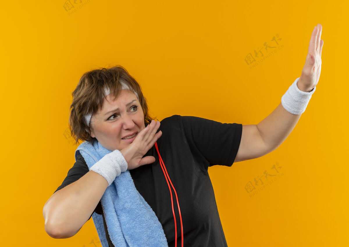 头带中年运动女性 身穿黑色t恤 头上戴着头巾 肩上搭着毛巾 站在橙色的墙上 做出一种带着恐惧表情的防御姿态中年手势表情