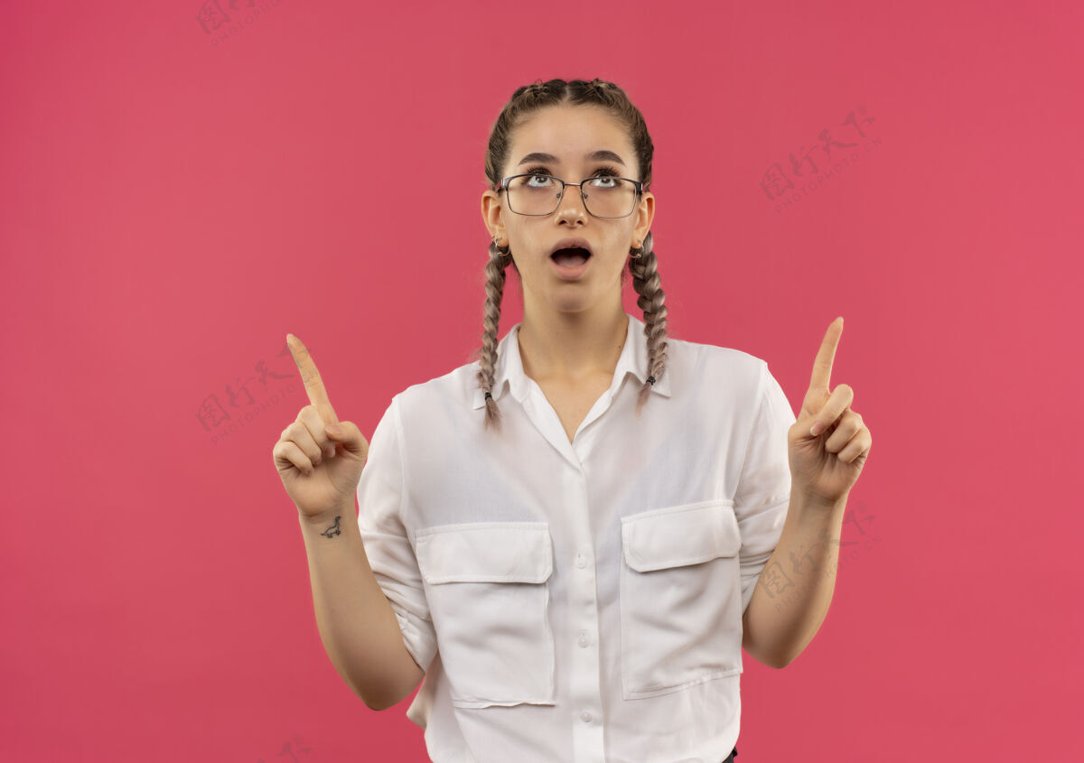 手指戴着眼镜 梳着辫子 穿着白衬衫的年轻女学生 食指朝上 站在粉红色的墙上 看上去很惊讶人指着辫子