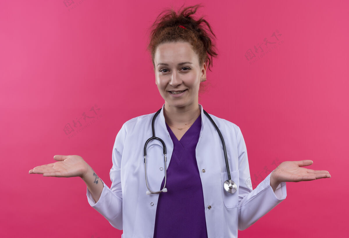 摊开年轻的女医生身穿白色外套 手持听诊器 微笑着张开双臂站在粉红色的墙上女人听诊器微笑