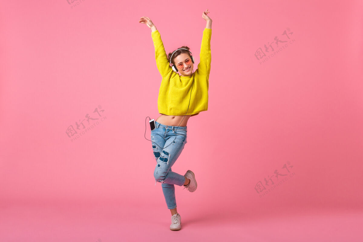 女孩迷人的快乐搞笑的女人穿着黄色毛衣跳舞戴着耳机听音乐穿着时髦的五颜六色的服装隔离在粉色的墙上 玩得开心声音明亮女人