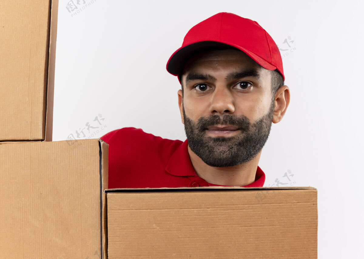 箱子留着胡须的送货员穿着红色制服 戴着一顶带纸板箱的帽子 站在白色的墙上偷看帽子偷看制服