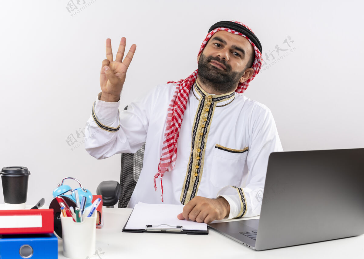 指指点点身着传统服装的阿拉伯商人坐在桌旁 手提电脑微笑着 手指朝上 三号手指在办公室工作工作阿拉伯语微笑