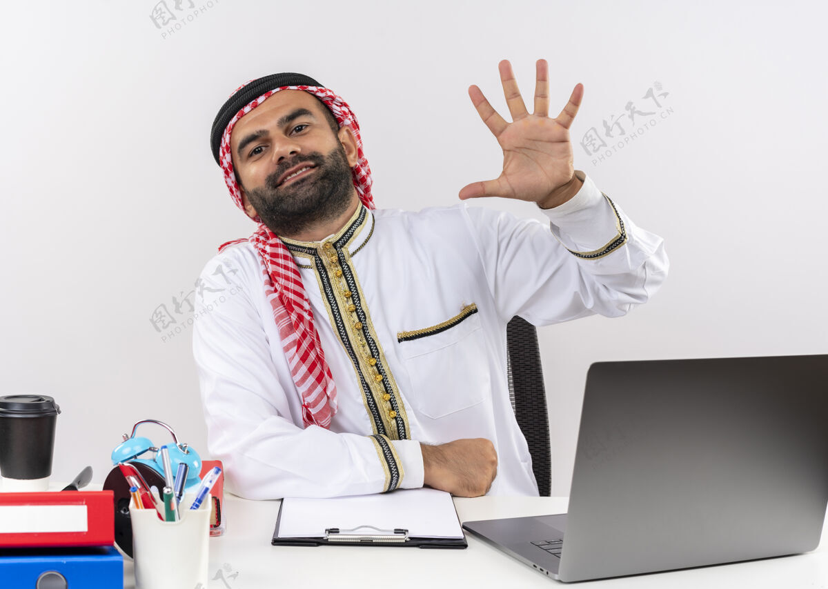 电脑身着传统服装的阿拉伯商人坐在桌旁 手提电脑在展示 手指朝上 5号微笑着在办公室工作传统五数字