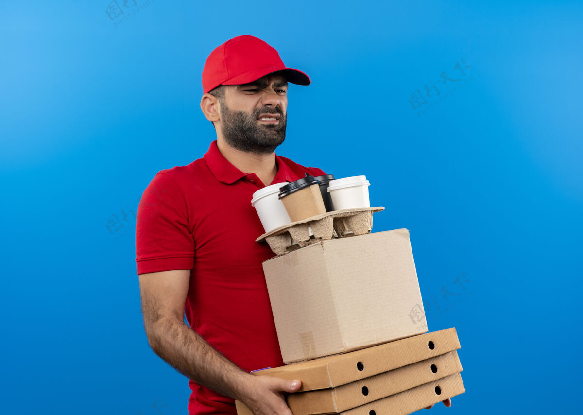 胡子留着胡子的送货员穿着红色制服 戴着帽子 手里拿着纸板箱 站在蓝色的墙上 不高兴地看着疲惫和过度劳累的他累了男人帽子