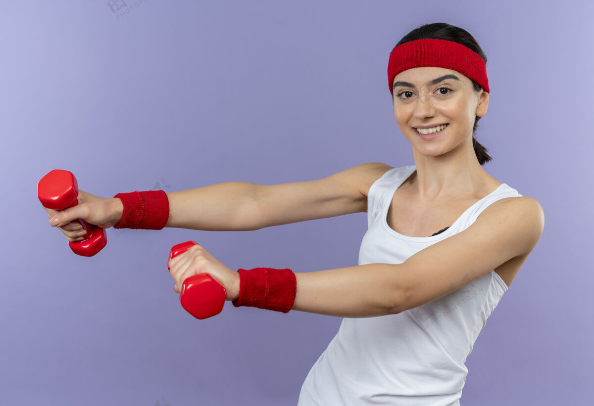 锻炼身穿运动服 头箍 双手举着两个哑铃 面带微笑的年轻健身女孩站在紫色的墙上做运动健身开心年轻