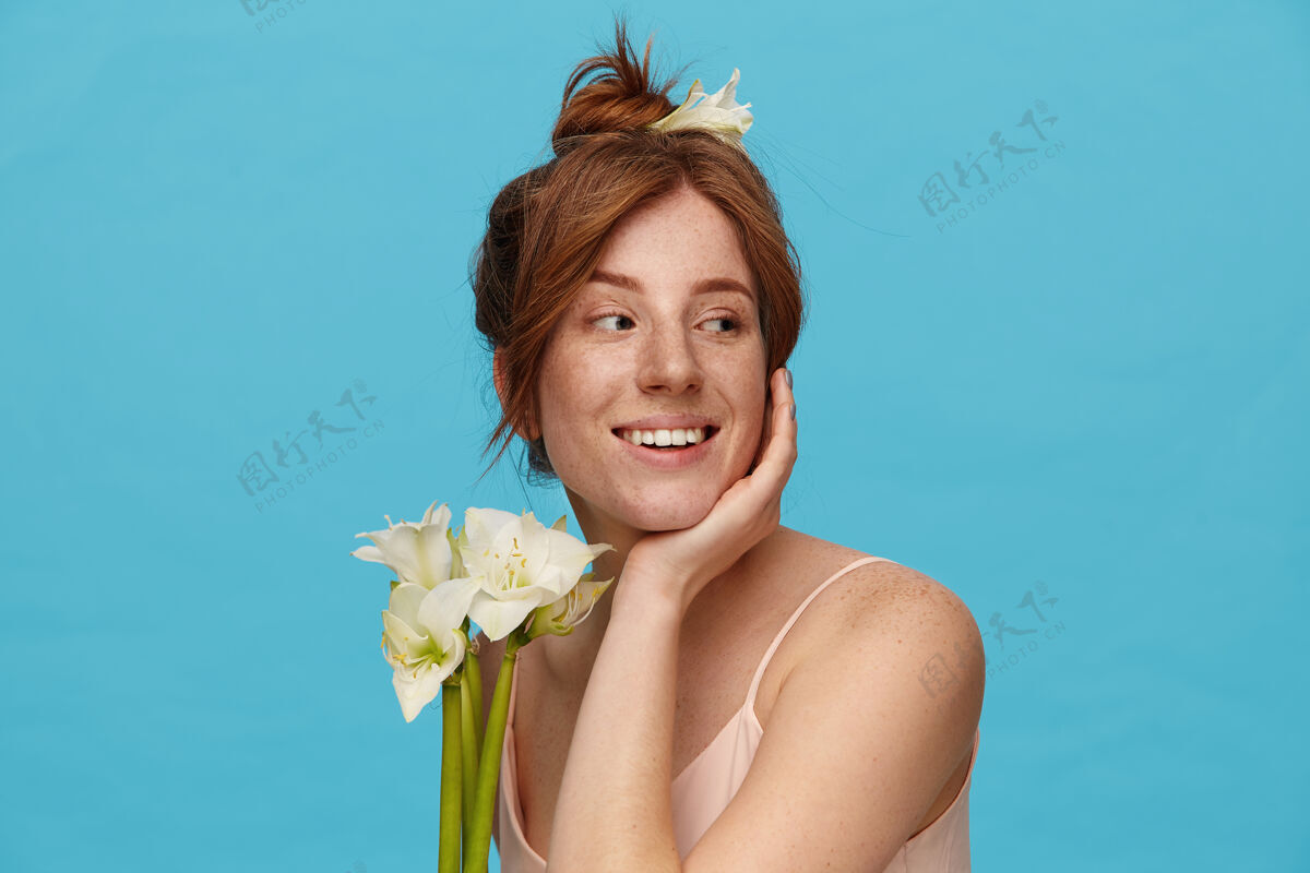 表情快乐的年轻可爱的红发女性 把头靠在举起的手上 带着愉快的微笑积极地看着一边 站在蓝色的背景上 鲜花盛开皮带白色年轻