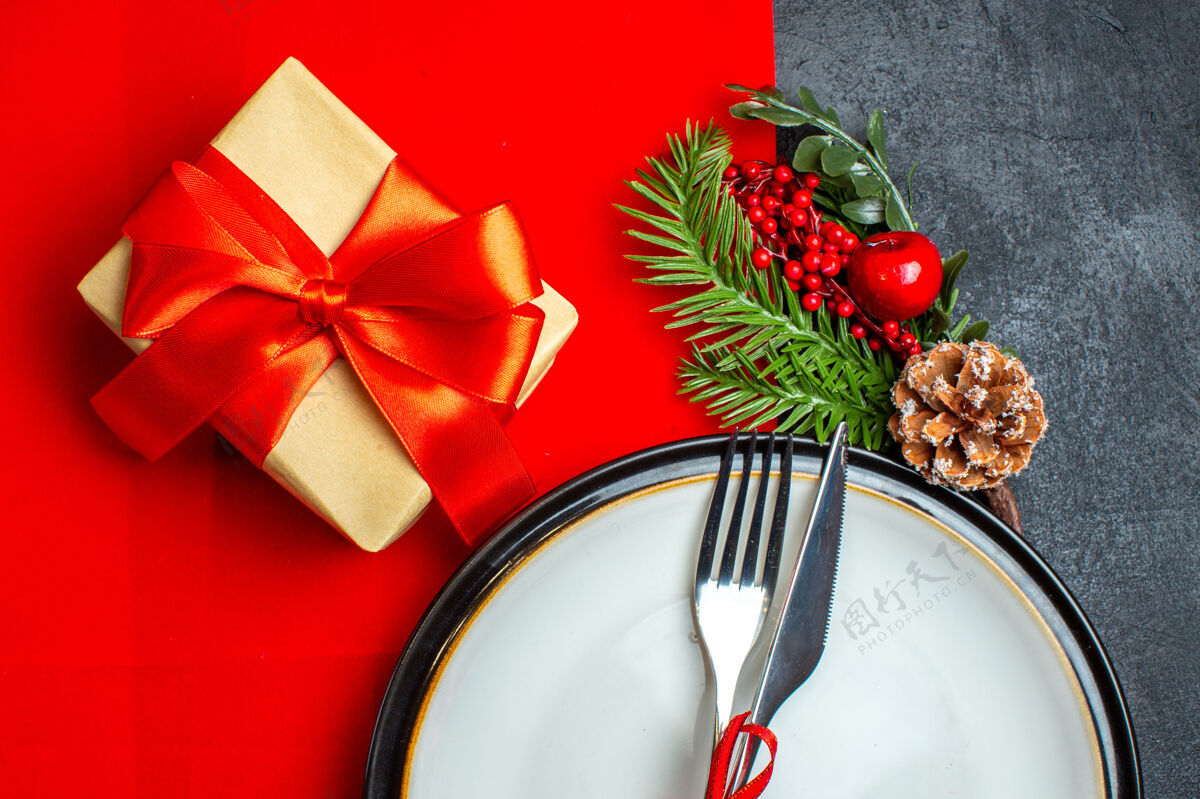 冬青新年背景的半张照片 餐具上镶着红丝带 餐盘上装饰着配件杉木树枝 旁边是一张红色餐巾上的礼物冷杉餐具一半