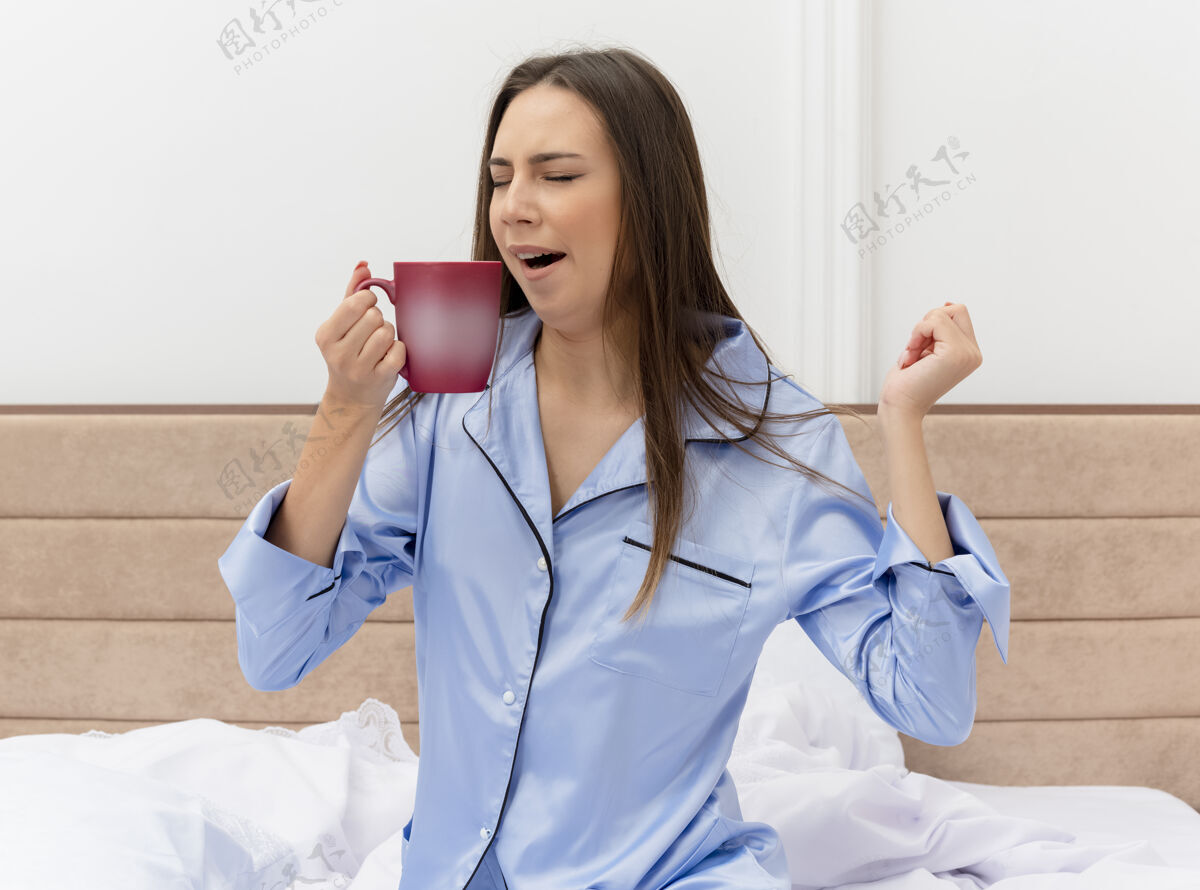 漂亮穿着蓝色睡衣的年轻漂亮女人坐在床上 端着一杯咖啡 醒来时在卧室里打着哈欠打哈欠坐着床