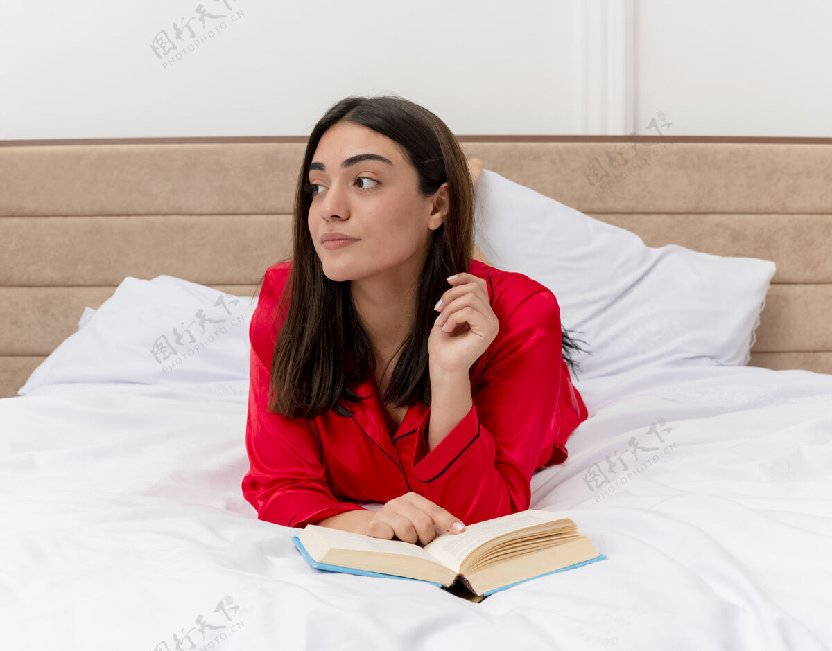 卧室穿着红色睡衣的年轻美女躺在床上 拿着书 悠闲地享受着周末 在卧室的灯光背景下 一边沉思着一边看沉思女人睡衣