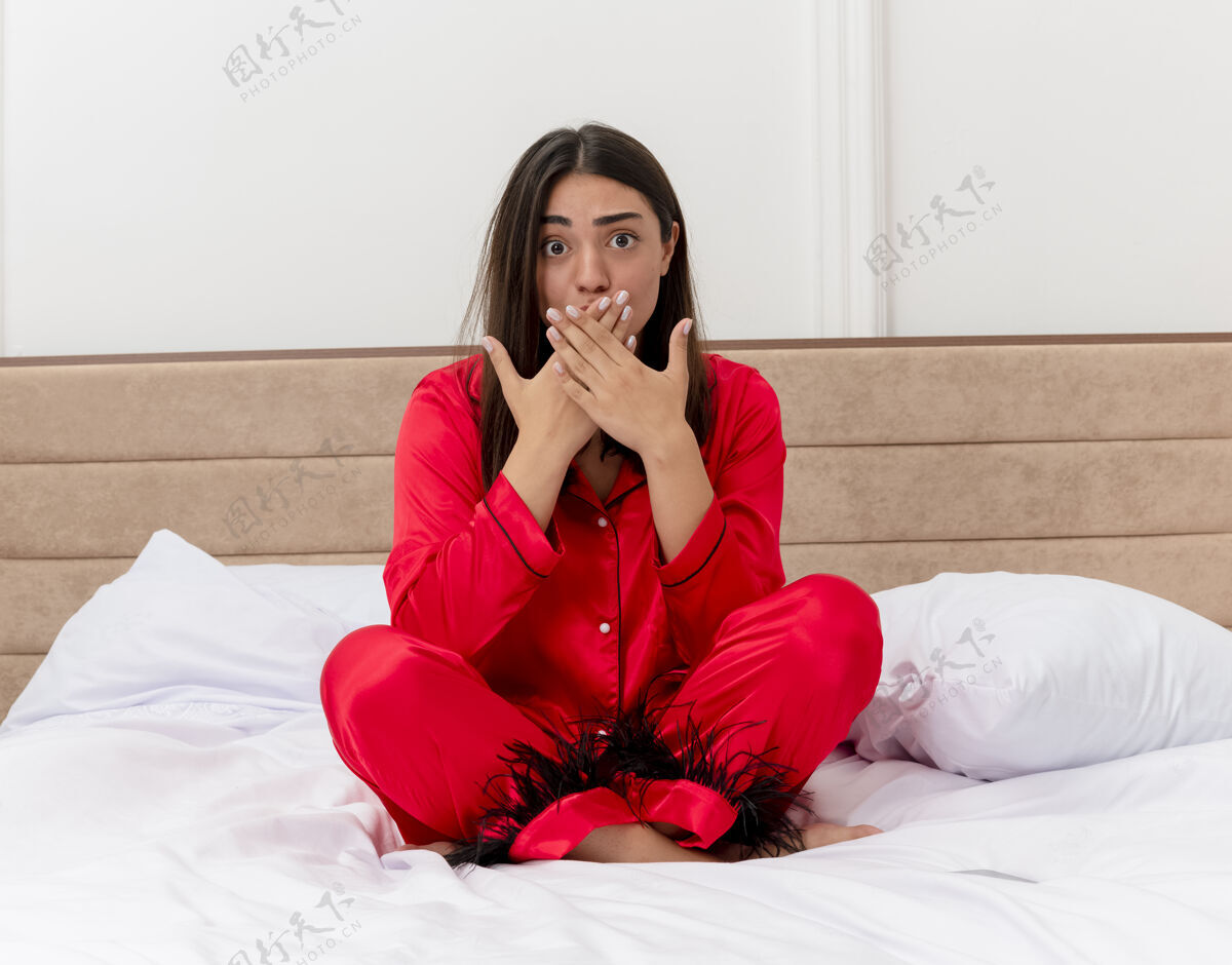 卧室穿着红色睡衣的年轻美女坐在床上看着摄像机 惊讶地用手捂住嘴巴 在卧室的室内灯光背景下手看年轻