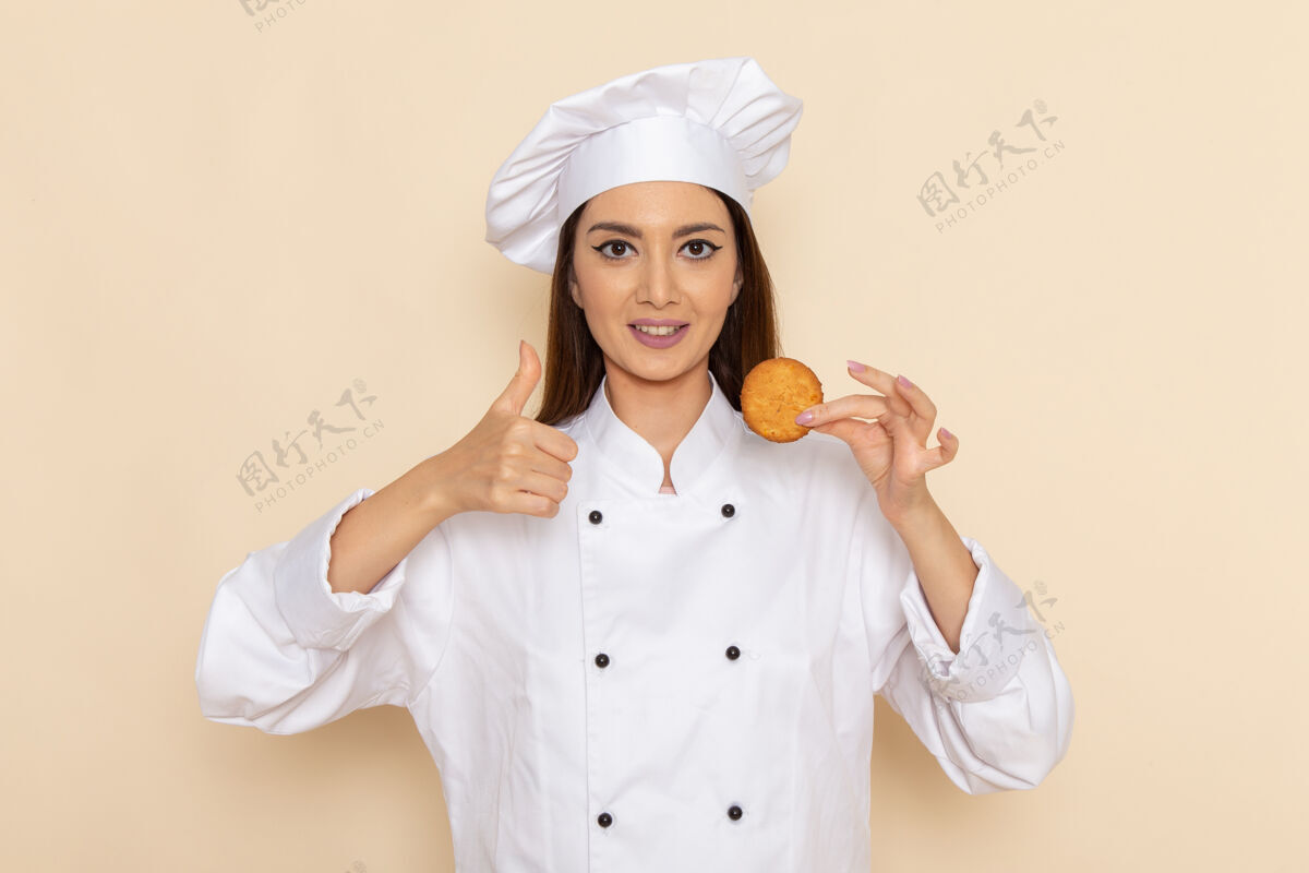 成人身穿白色厨师服的年轻女厨师正拿着小饼干站在浅白的墙上厨房前面人