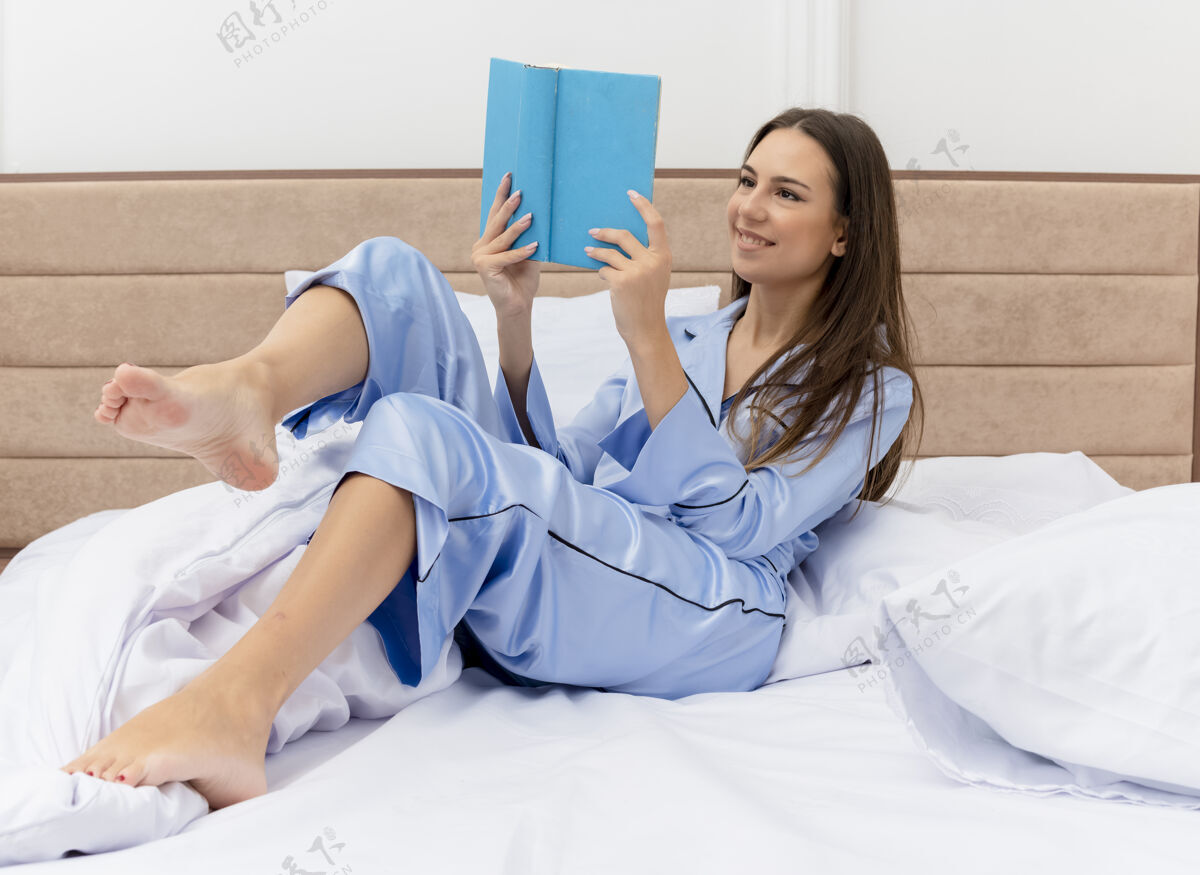 快乐穿着蓝色睡衣的年轻美女躺在床上看书 面带笑容 在卧室室内灯光背景下享受周末卧室周末美丽
