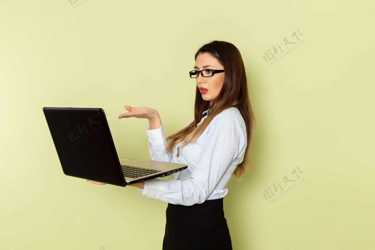 便携式穿着白衬衫和黑裙子的女上班族正拿着笔记本电脑站在绿色的墙上前面女工作