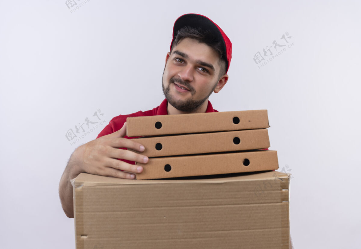 男人年轻的送货员穿着红色制服 戴着帽子 手里拿着一叠比萨饼盒和纸板盒 友好地微笑着看着镜头制服红色披萨
