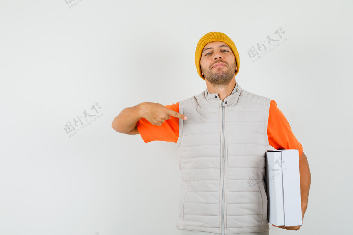 肖像一个穿着t恤 夹克 帽子的年轻人指着纸板箱 看上去很自豪开朗盒子休闲